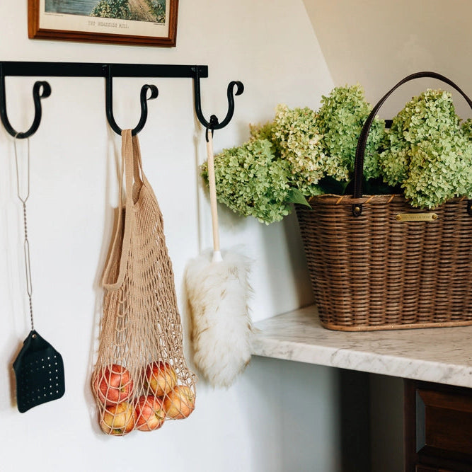 Hanging hooks with a net bag, broom, and duster against a wall with a basket of flowers on a shelf.