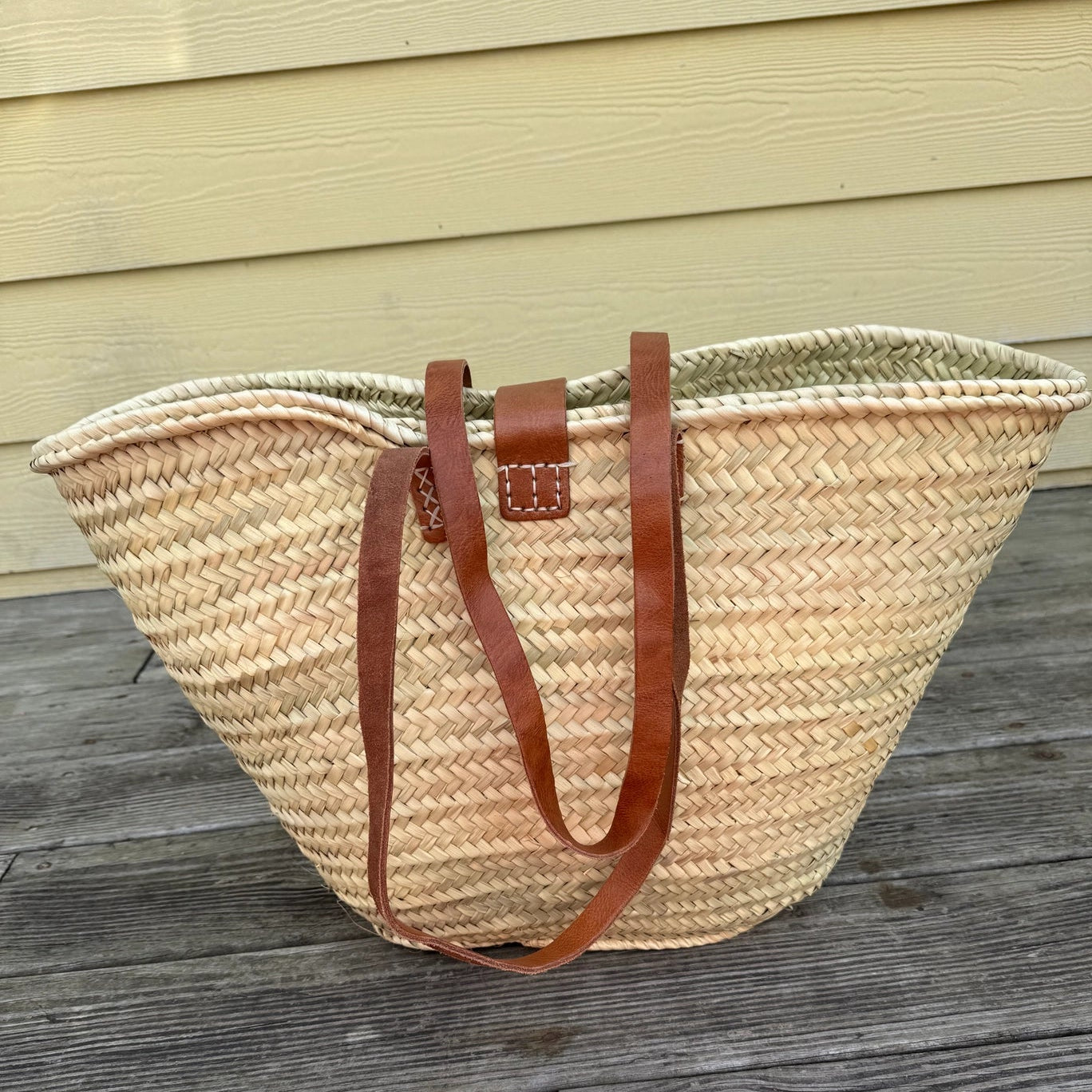A large woven straw tote bag with brown leather shoulder straps, resting on a weathered gray wooden deck in front of a light yellow house siding background.