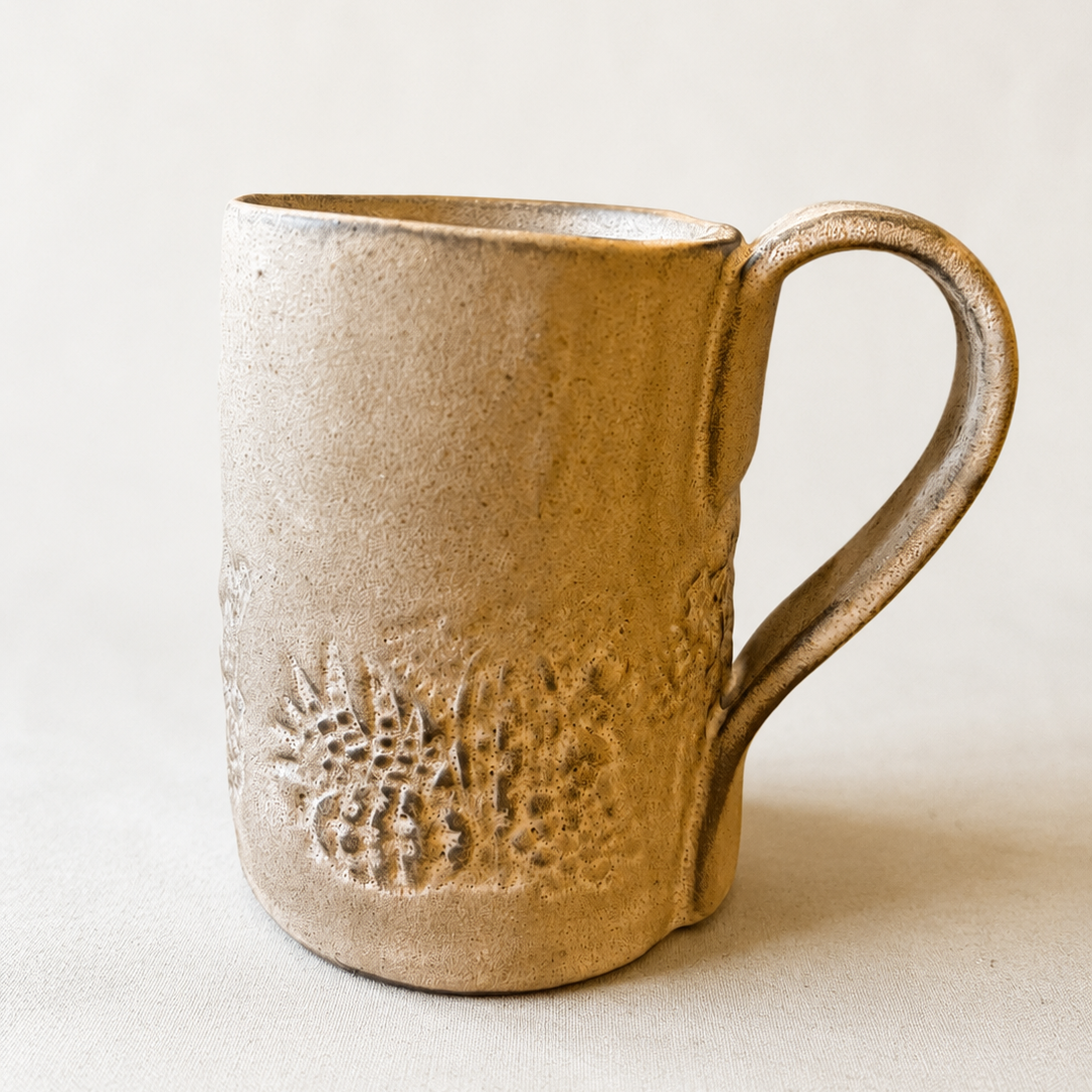 A handmade ceramic mug with a textured, organic shape and embossed floral pattern near the base, shown on a neutral beige background.