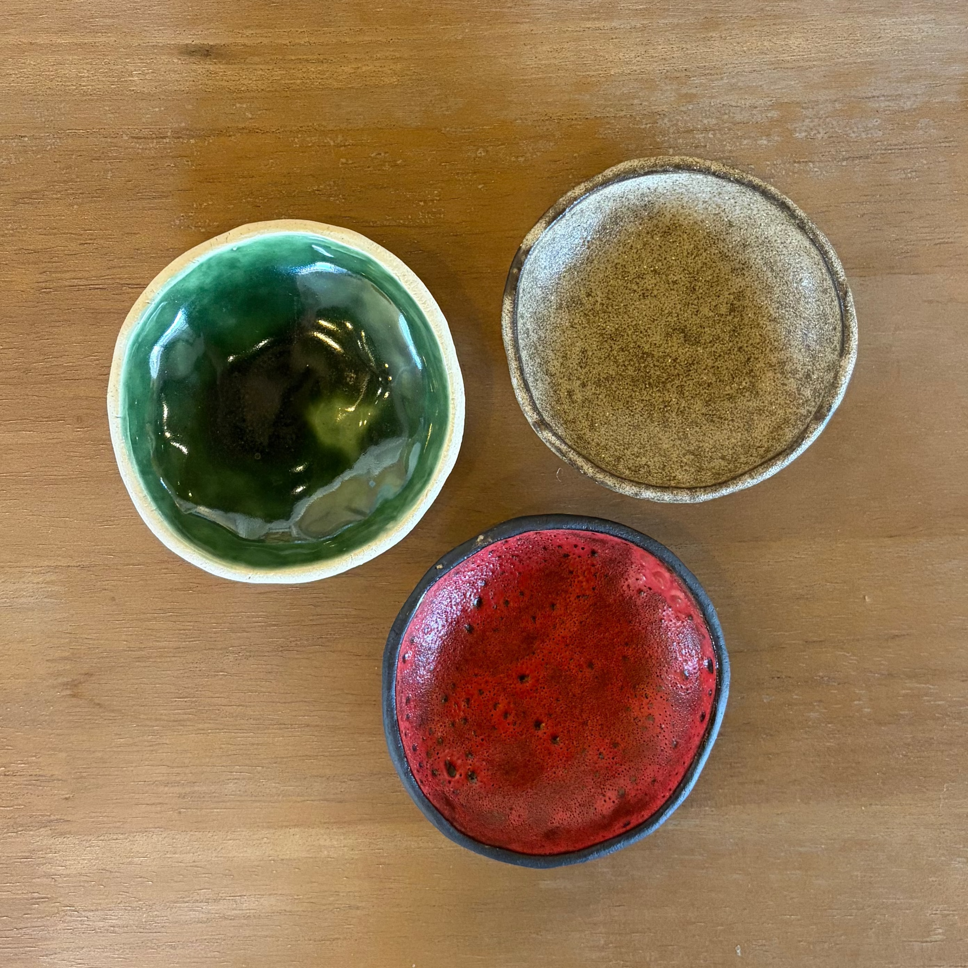 Three small handmade ceramic bowls in green, beige, and red glazes arranged on a wooden serving board on a dark wood table.