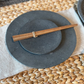 A hand-carved, round, gray stoneshard plate with a wooden stylus on top, placed on a woven table mat.