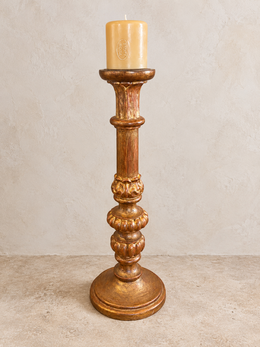 A tall, ornate wooden candlestick holder with a carved, distressed finish in warm brown and gold tones, holding a cream-colored pillar candle, set against a textured beige wall and floor.