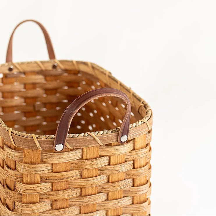 A close-up of a woven natural fiber basket with dark brown leather handles and silver metal rivets, set against a plain white background.