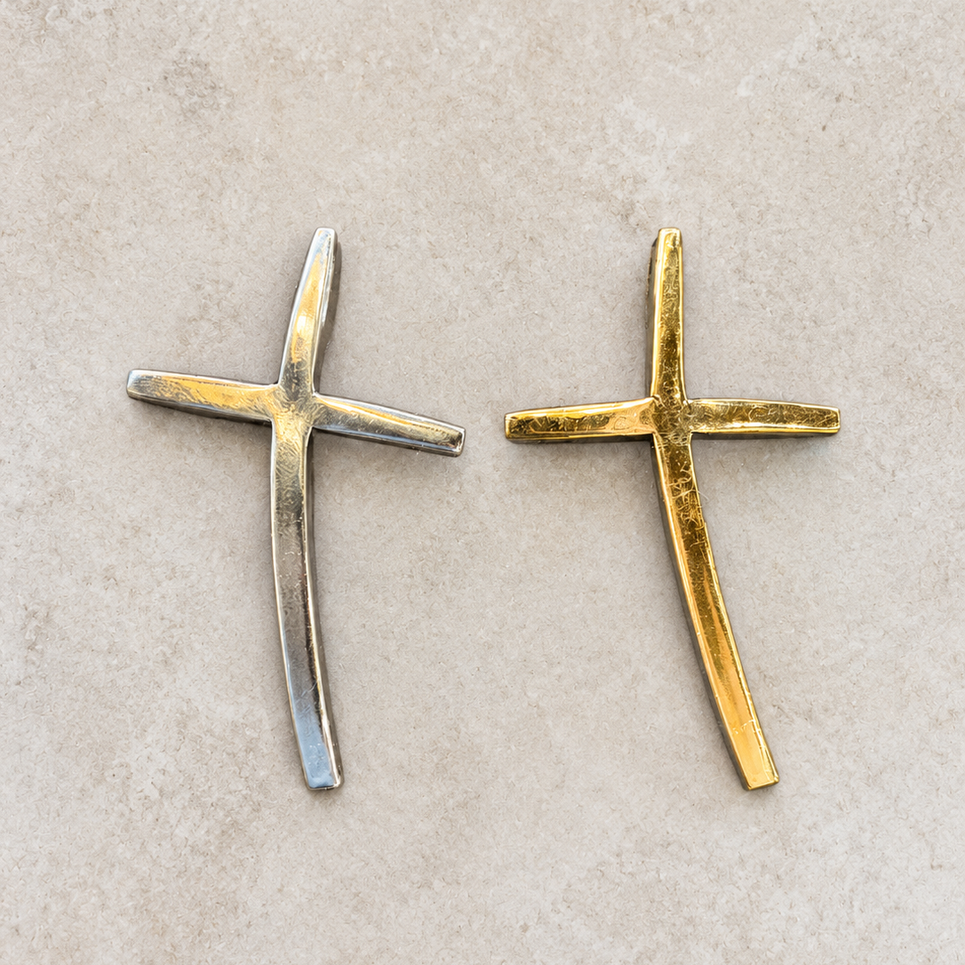 Two small cross-shaped jewelry pieces, one silver-toned and one gold-toned, displayed side by side on a light beige textured surface.