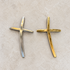 Two small cross-shaped jewelry pieces, one silver-toned and one gold-toned, displayed side by side on a light beige textured surface.