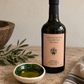 A bottle of Heritage Extra Virgin Olive Oil stands on a stone surface next to a small bowl of oil, olive branches, and a woven cloth. The bottle has a black cap and a peach label reading 'California Harvest from 150 Year Old Trees'.