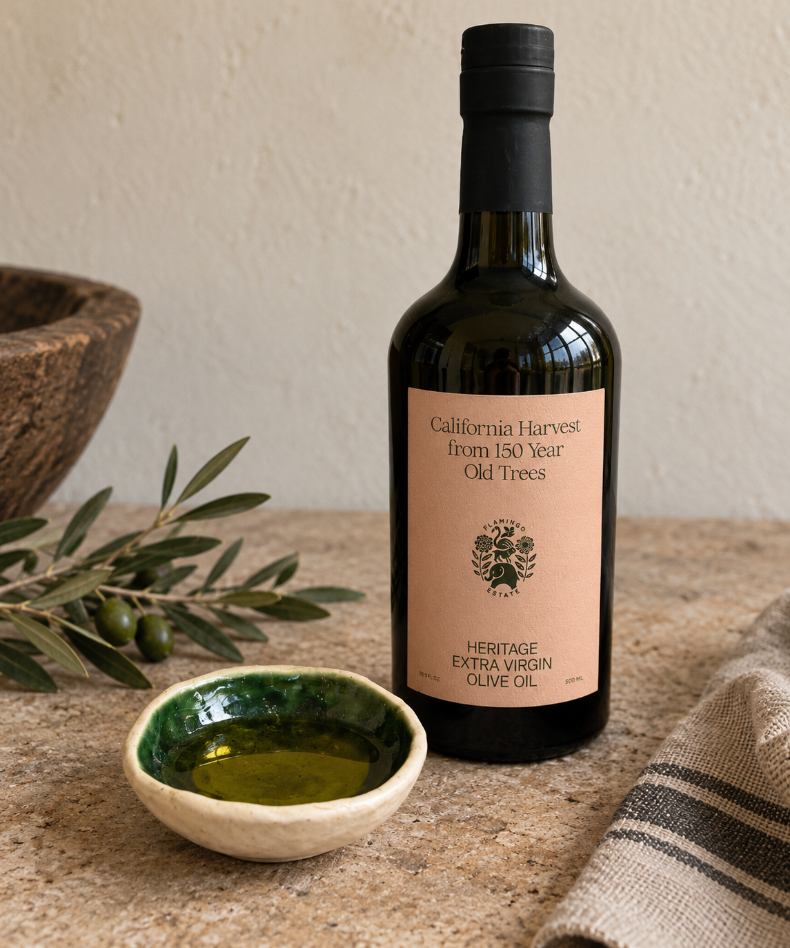 A bottle of Heritage Extra Virgin Olive Oil stands on a stone surface next to a small bowl of oil, olive branches, and a woven cloth. The bottle has a black cap and a peach label reading 'California Harvest from 150 Year Old Trees'.