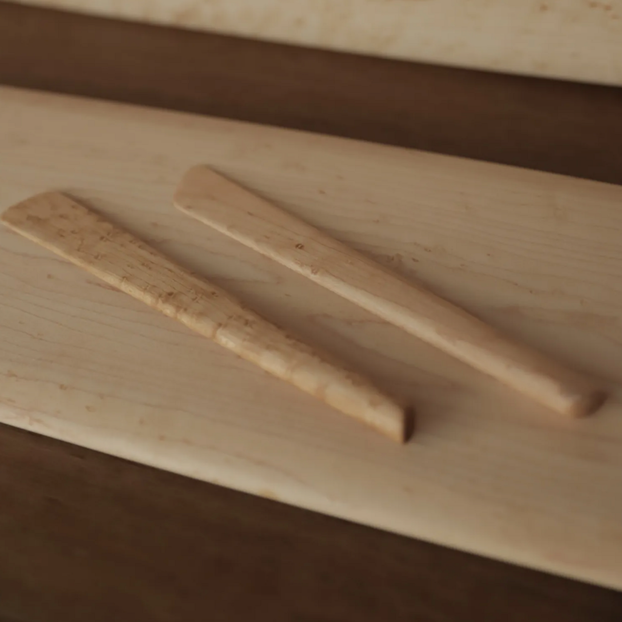 Two wooden cheese knives with bird's-eye maple pattern lying on a light wooden surface.